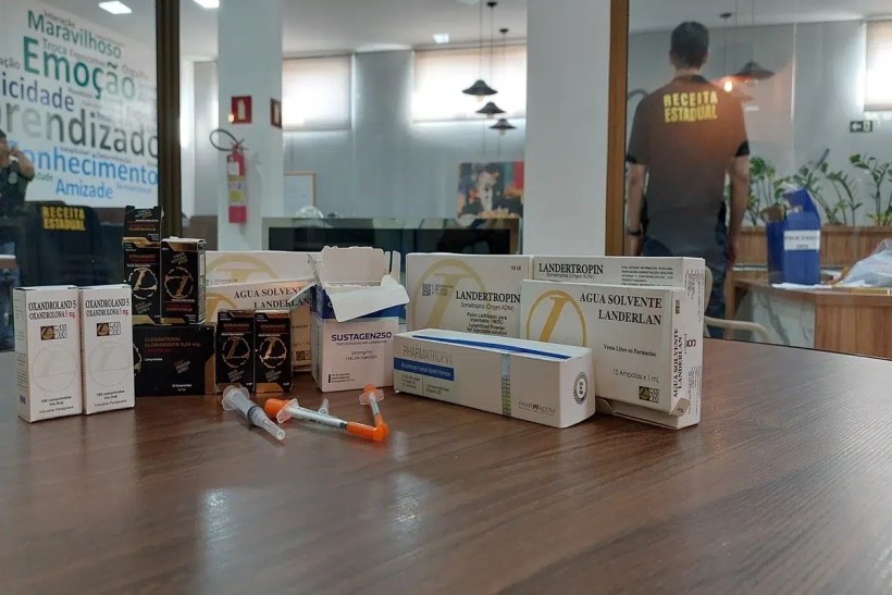 Vários medicamentos e seringas dispostos sobre uma mesa em um ambiente de escritório, com um fundo que exibe uma parede decorada com palavras relacionadas a emoção e aprendizado.