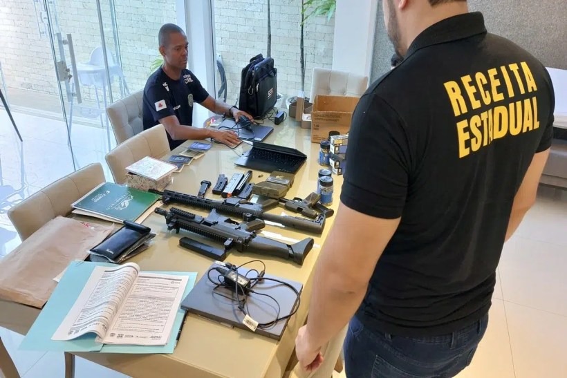 Agentes em uma sala analisando armas e documentos em uma mesa. Um agente está sentado com um laptop, enquanto outro está em pé, usando uma camisa com a inscrição 'Receita Estadual'.