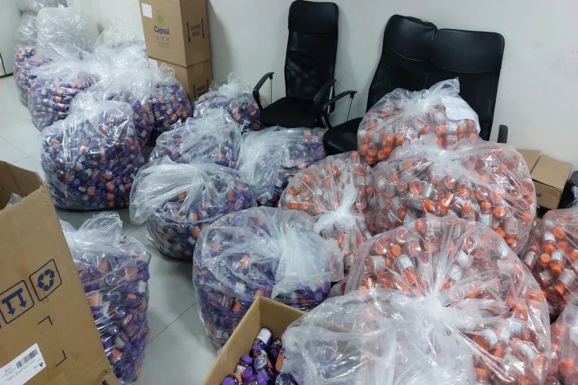 Várias sacolas plásticas cheias de frascos coloridos, com duas cadeiras pretas ao fundo e caixas de papelão.