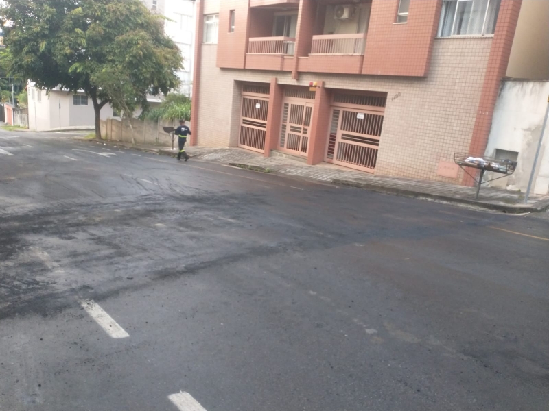 Rua residencial com asfalto escuro, mostrando um homem caminhando e edifícios ao fundo, incluindo garagens.