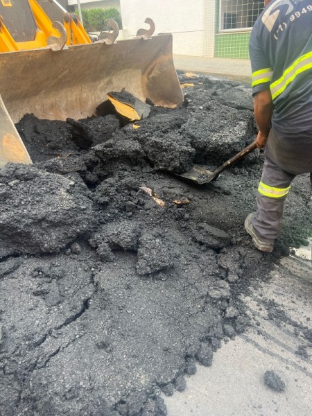 Homem trabalhando com uma pá, removendo asfalto de uma pista em obras, com uma retroescavadeira ao fundo.