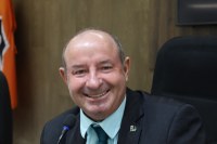 Homem sorrindo em ambiente formal, usando terno escuro e gravata, com microfone à frente.