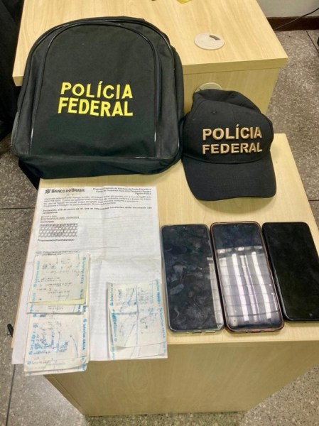 Mochila e boné da Polícia Federal, um documento bancário e três smartphones sobre uma mesa.