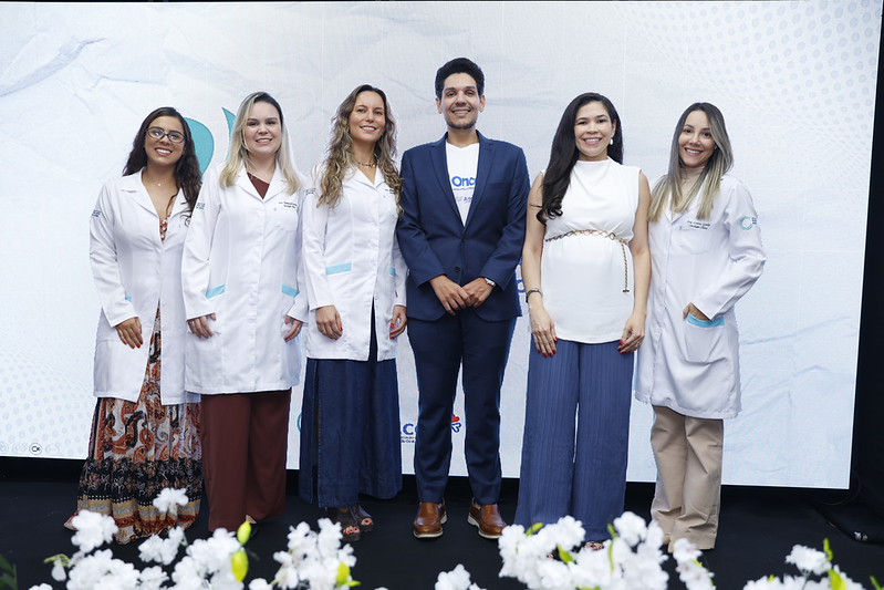 Seis profissionais de saúde, incluindo médicos e enfermeiras, posam para a foto em um evento. Eles estão vestidos com jalecos brancos e roupas casuais, sorrindo em frente a um fundo branco com elementos gráficos.