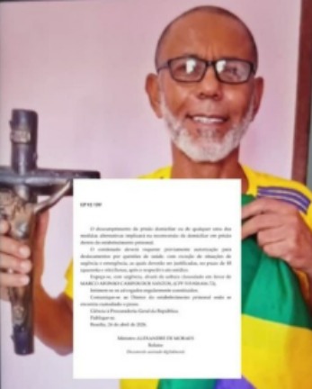 Homem sorridente usando óculos, segurando uma cruz e uma bandeira do Brasil, com um documento em destaque ao fundo.