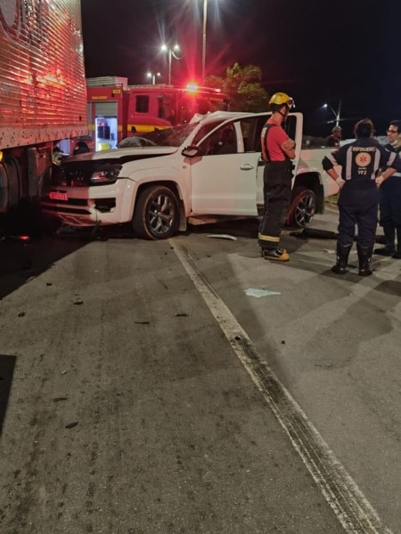 Acidente de trânsito à noite envolvendo um carro branco e um caminhão, com membros dos serviços de emergência ao redor.