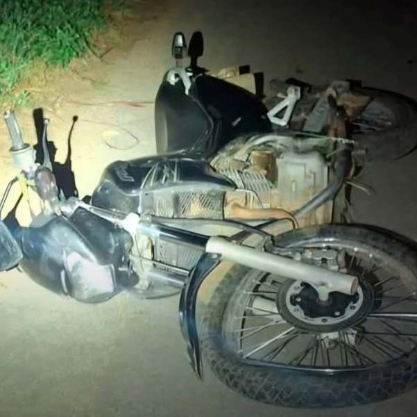 Motocicleta tombada no chão, com rodas para cima e visível dano ao corpo da moto, em um ambiente escuro.