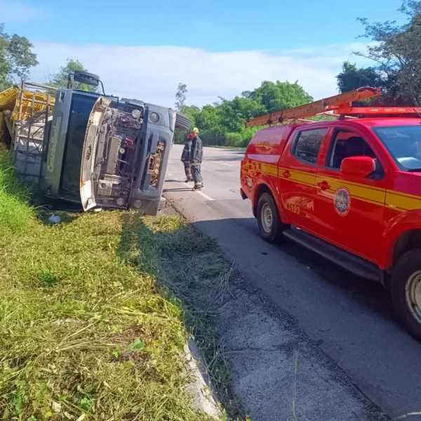 Caminhão tombado ao lado da estrada, com um veículo de bombeiros vermelho próximo e bombeiros ao fundo.