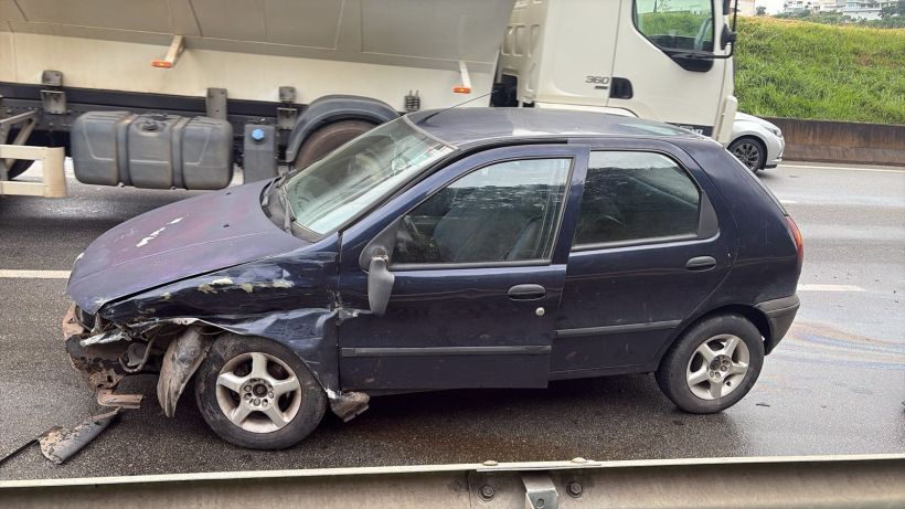 Carro preto danificado na estrada, com a frente amassada e danificada, estacionado próximo a um caminhão.