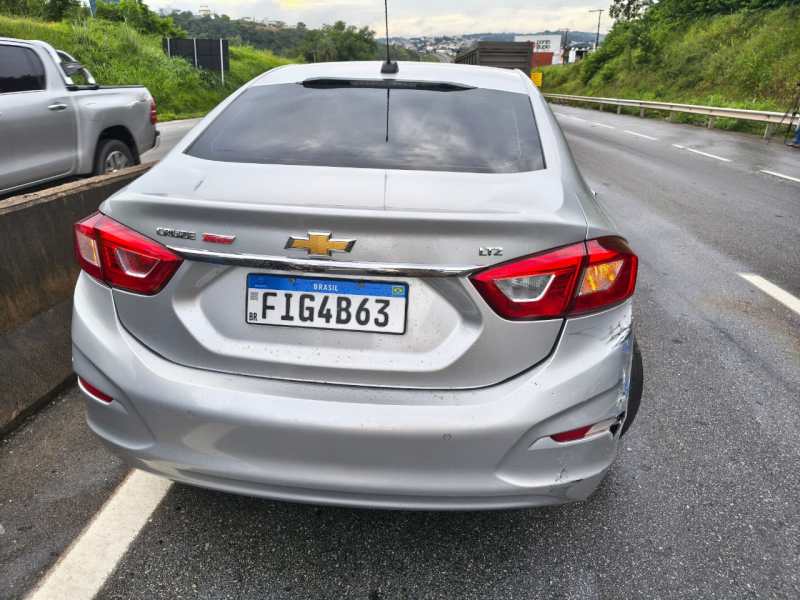 Imagem traseira de um carro Chevrolet Cruze prata, com danos visíveis na lateral direita. O veículo está estacionado próximo a uma estrada.