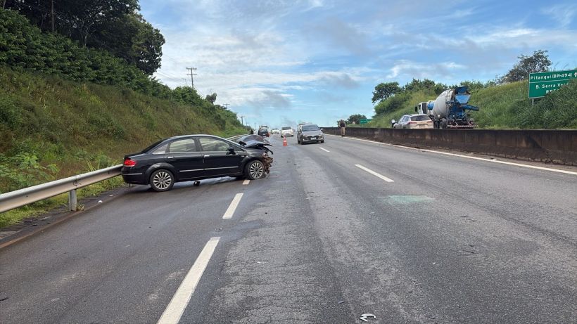 Imagem de uma estrada com um carro preto acidentado parado no acostamento. Há outros veículos em movimento ao fundo e uma placa de sinalização à direita.