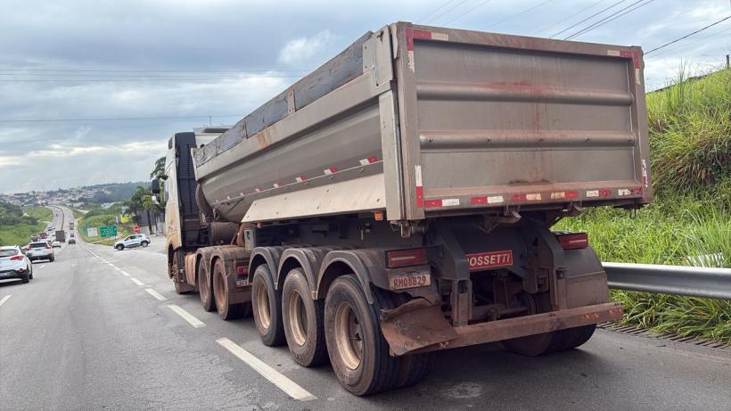 Caminhão basculante estacionado na beira da estrada, com uma estrada movimentada ao fundo e vegetação à direita.