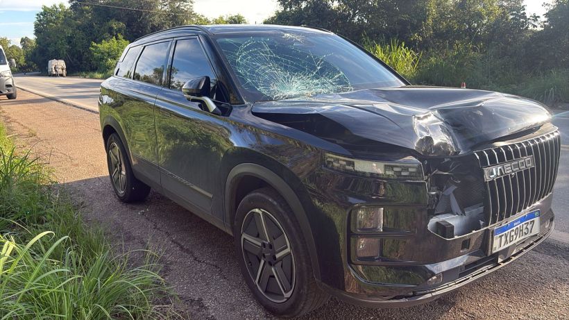 Veículo SUV preto com danos na parte frontal e para-brisa quebrado, estacionado à beira da estrada em uma área verde.