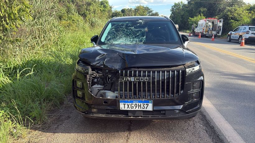 Veículo SUV preto com o capô amassado e para-brisa quebrado, estacionado em uma estrada com vegetação ao redor.