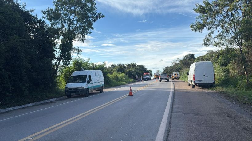 Uma estrada com veículos estacionados à beira, sinalização com cone e vegetação ao redor.