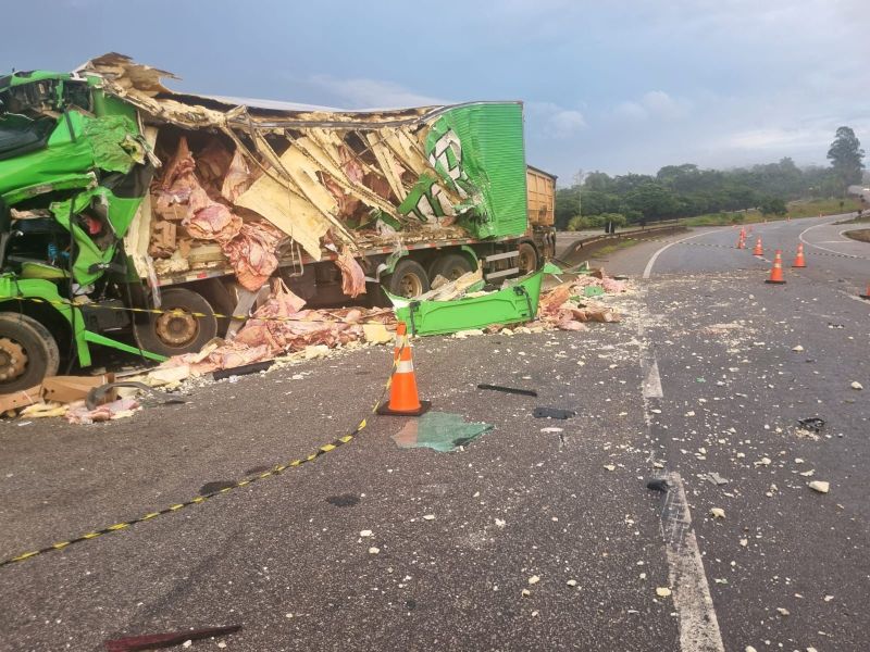 Caminhão acidentado na estrada, com carga espalhada e danos visíveis na carroceria e no asfalto, cercado por cones laranjas e fita de contenção.