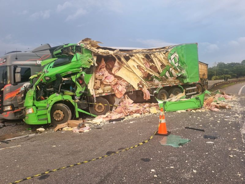 Acidente envolvendo caminhão verde na estrada, com a cabine destruída e carga espalhada pelo asfalto.