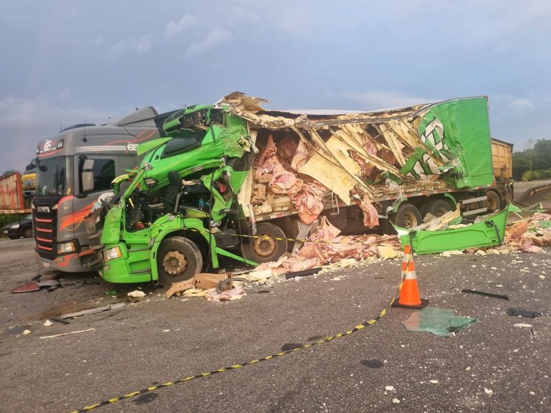 Caminhões envoltos em um acidente, com a cabine de um caminhão verde danificada e carga espalhada pelo chão.