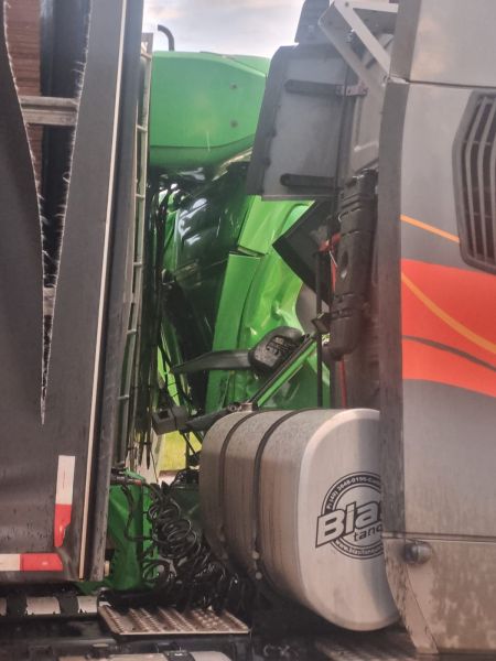 Imagem de um caminhão com um reboque, mostrando parte verde de um veículo agrícola preso na lateral.