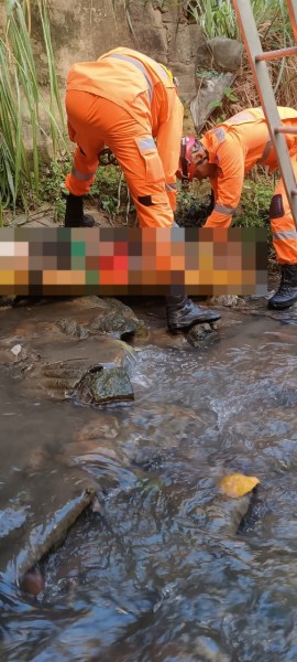 Dois profissionais em trajes de segurança laranja examinando uma área ao lado de um rio, com pedras visíveis na água.