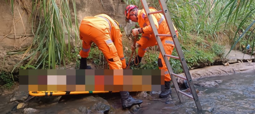 Dois socorristas em trajes laranja trabalham na beira de um rio, preparando a remoção de uma maca com um corpo, cercados por vegetação.