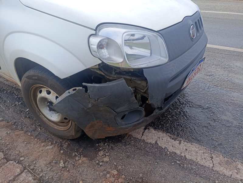 Carro branco com o para-choque frontal danificado na lateral direita, exposto e parcialmente quebrado, estacionado em uma estrada.