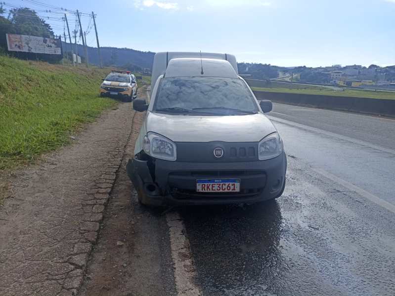 Furgão cinza estacionado com danos na frente, à beira da estrada, com um carro da polícia visível ao fundo.