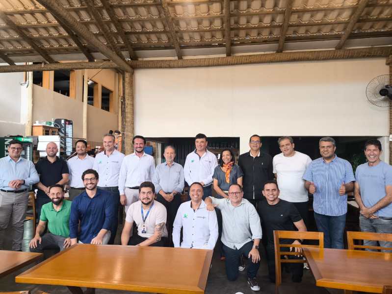 Um grupo de pessoas posando em um restaurante, com mesas de madeira ao fundo. Algumas pessoas estão de pé e outras sentadas, todas sorrindo e fazendo gestos de positivo.