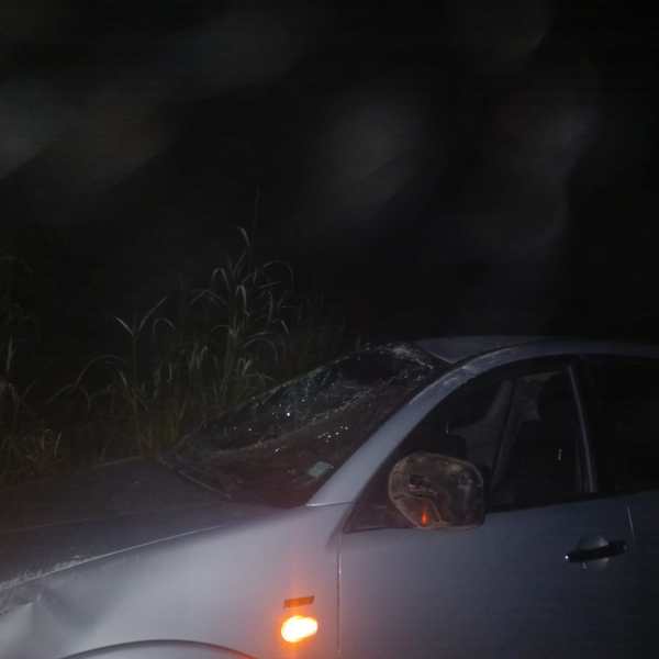 Carro danificado à noite, com o para-brisa quebrado e vegetação ao fundo.
