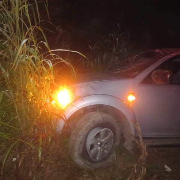 Carro acidentado em área de vegetação, com farol ligado, à noite.