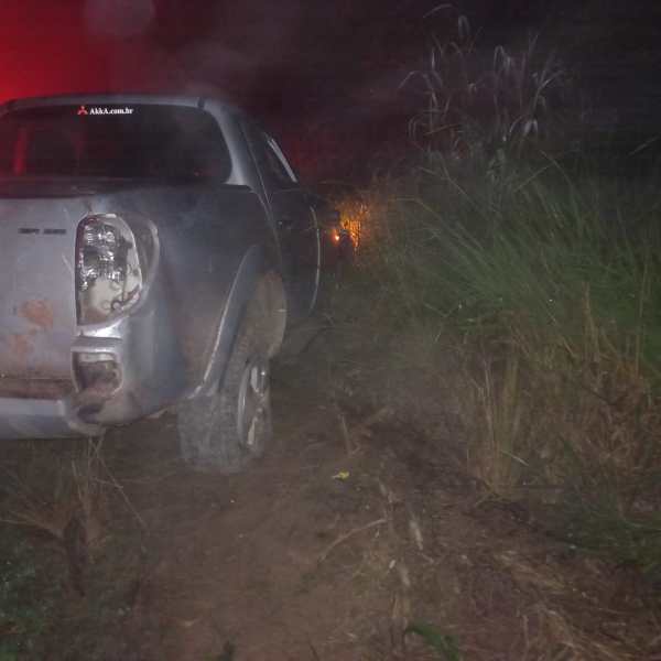 Carro parado em um caminho estreito, com vegetação ao redor e luzes ao fundo, durante a noite.