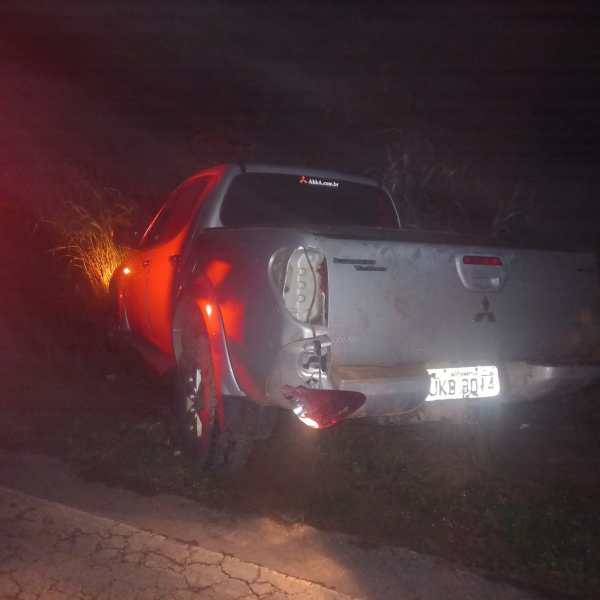 Um carro pick-up Mitsubishi prateado estacionado em uma área escura, com danos visíveis na parte traseira e luzes de posição acesas.