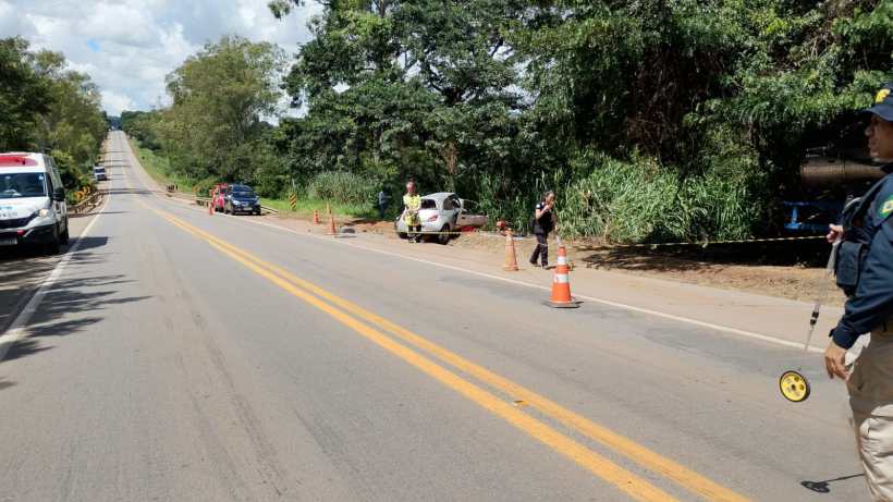 Cena de uma estrada com carros, incluindo ambulância, e cones de sinalização, onde policiais estão realizando um trabalho de investigação ao lado de um veículo parado.