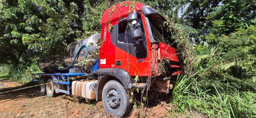 Caminhão vermelho parcialmente coberto por vegetação, estacionado em uma área rural com árvores e grama ao redor.