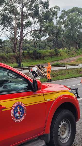 Veículo acidentado na margem da estrada com um bombeiro em equipamento de segurança laranja ao lado, enquanto um caminhão do Corpo de Bombeiros de Minas Gerais está em primeiro plano.