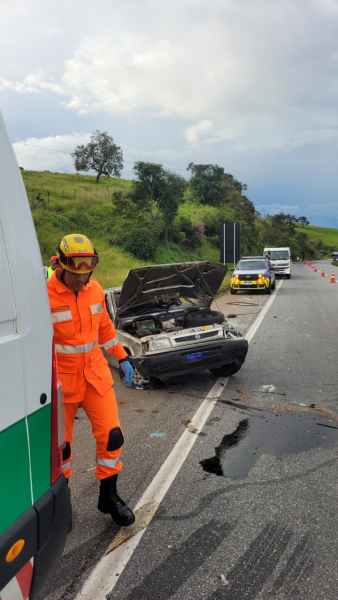 Um trabalhador de emergência vestido com um uniforme laranja e capacete, ao lado de um veículo acidentado com o capô aberto na estrada. Ao fundo, outros veículos estão parados e cones de sinalização estão visíveis.