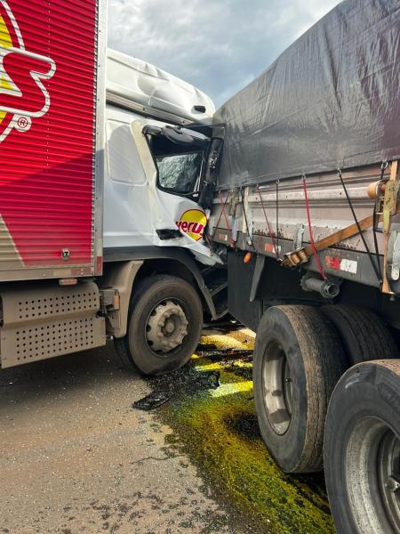 Colisão entre dois caminhões na estrada, com um deles parcialmente danificado e descarregando algum tipo de líquido no chão.
