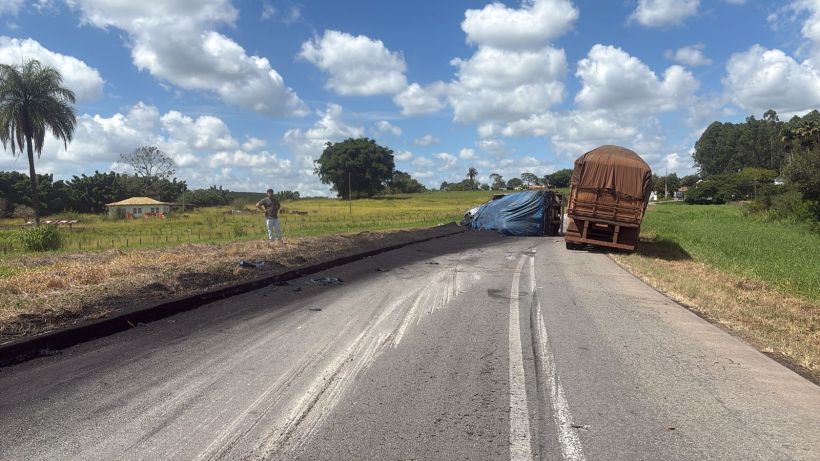 Caminhão tombado na estrada, com carga sobre a via e vegetação ao fundo. Um homem observa a cena ao lado.