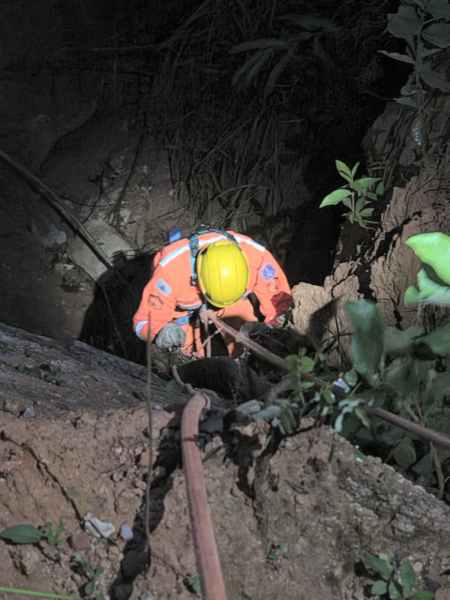 Um trabalhador de resgate usando um macacão laranja e um capacete amarelo está escorregando para baixo em um buraco na terra, rodeado por vegetação.
