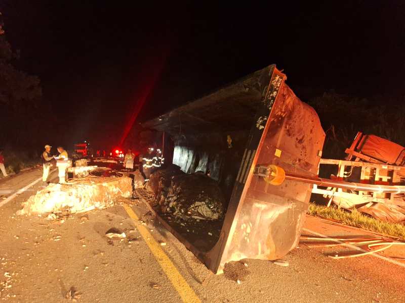 Caminhão tombado na estrada durante a noite, com equipes de resgate trabalhando ao redor.