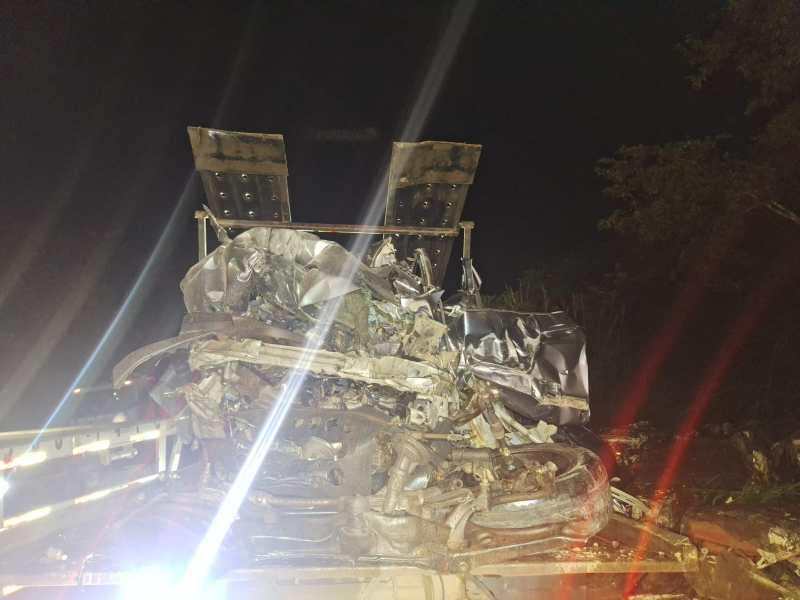Acidente de carro à noite, com um veículo severamente danificado, encostado em um caminhão.