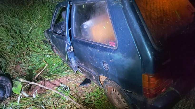 Um carro azul tombado em uma área coberta de grama alta, com uma porta aberta e visíveis danos na carroceria.
