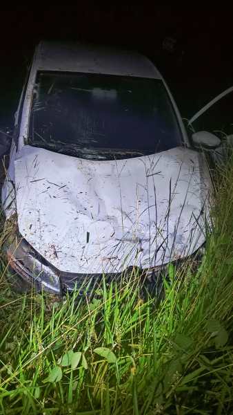 Um carro branco abandonado coberto de sujeira, situado em alta grama durante a noite.