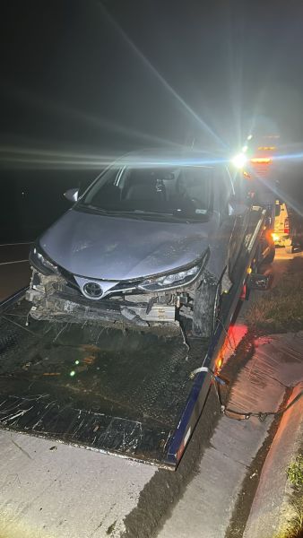 Carro acidentado sendo rebocado à noite, com danos visíveis na frente.