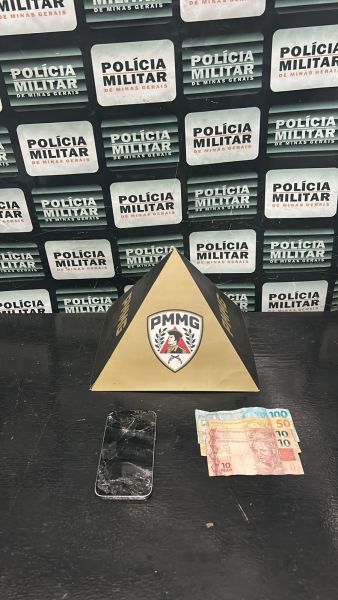 Um objeto em formato de pirâmide com o logotipo da PMMG, um celular preto e notas de dinheiro em uma superfície escura, com fundo de placas da Polícia Militar.
