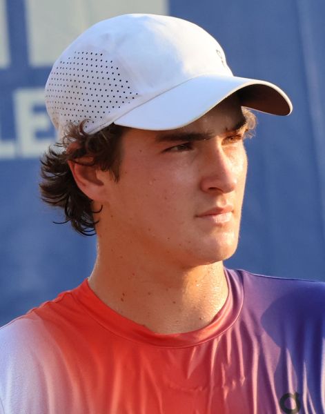 A young male tennis player wearing a white cap and an orange to purple gradient shirt, looking serious while standing outdoors.