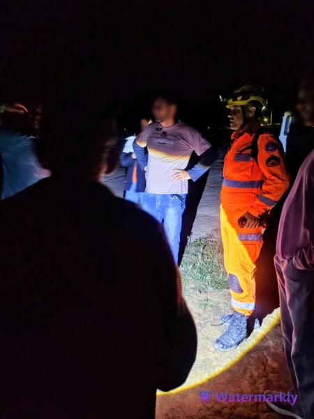 Grupo de pessoas interagindo à noite, com um profissional de resgate vestido de uniforme laranja e um capacete, observando a situação.