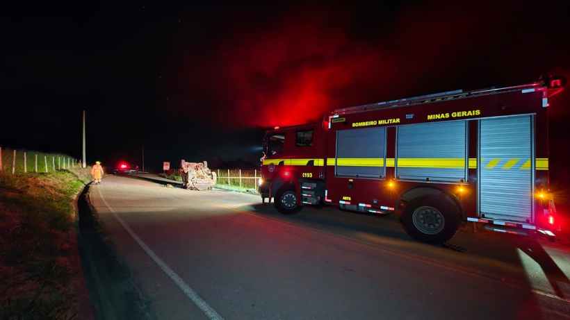 Um caminhão de bombeiros estacionado à beira da estrada durante a noite, com luzes vermelhas piscando e fumaça ao fundo.