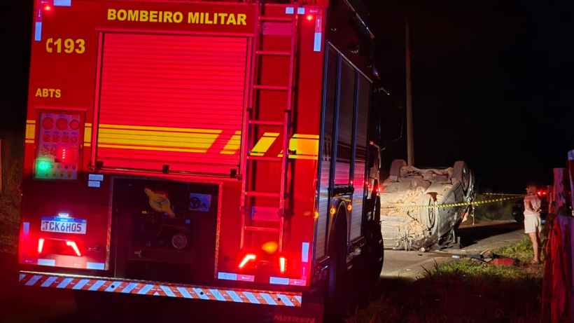Caminhão do Corpo de Bombeiros Militar com luzes vermelhas, próximo a um carro acidentado deitado na estrada, à noite.