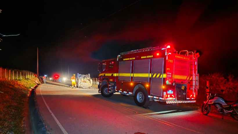 Caminho à noite com um caminhão de bombeiros vermelho estacionado, iluminação de emergência ativada, e uma cena de acidente ao fundo.